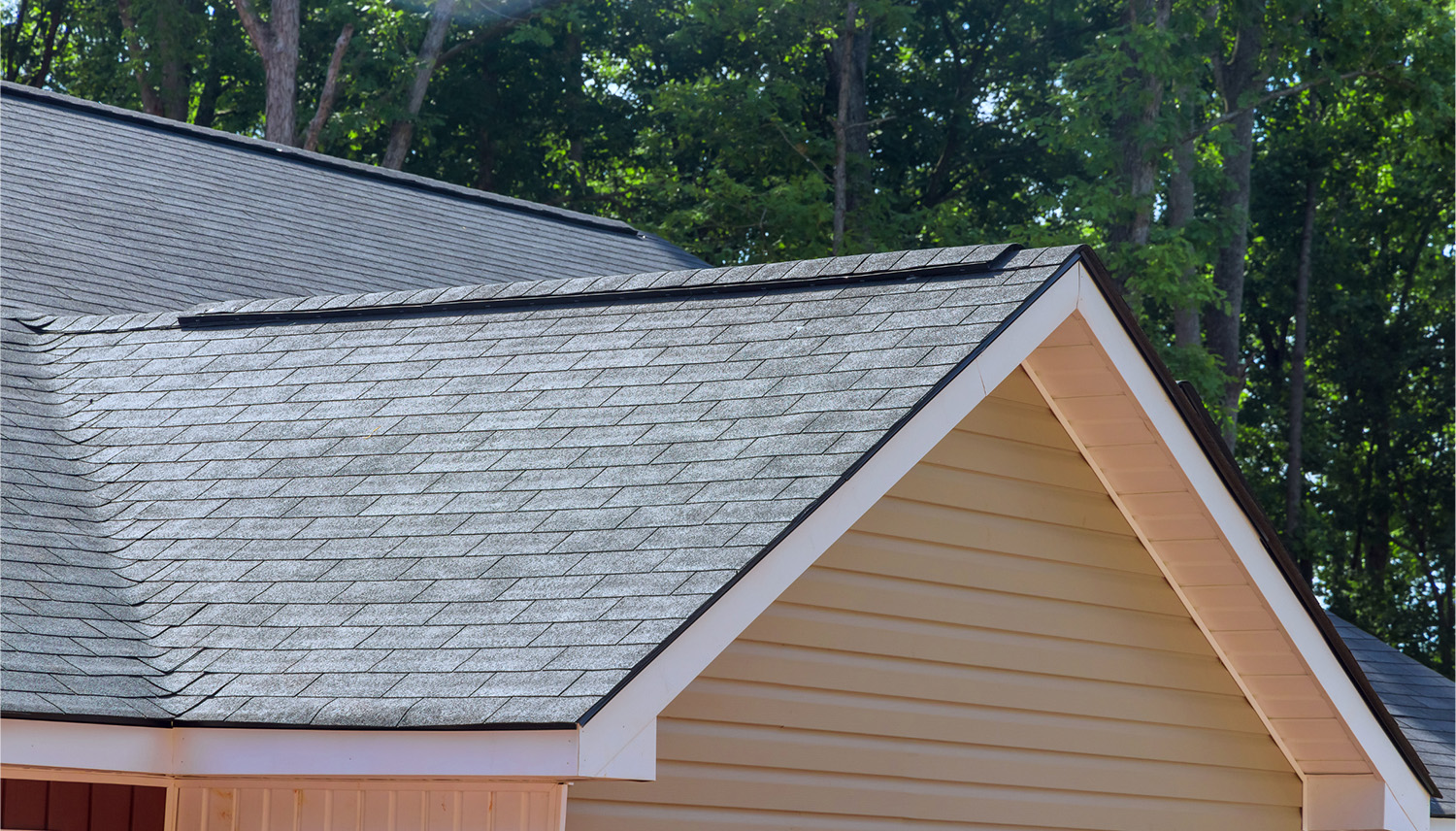 Close-up of a gray shingled roof with white trim and a triangular gable against a backdrop of green trees.